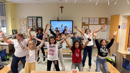 Eine Gruppe von Kindern in einem Klassenzimmer l&auml;chelt und streckt begeistert die Arme in die H&ouml;he.